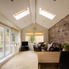 Cozy sunroom with a sloped ceiling, featuring skylights and large windows. A brick wall on the right, a TV, and leather chairs create a comfortable seating area. A wooden table and decorative items add warmth to the bright space.