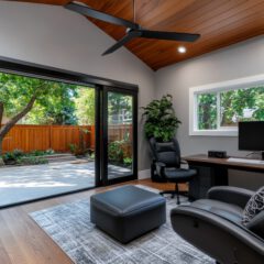 A modern home office with a desk, computer, and office chairs. Large glass doors open to a patio with trees and a wooden fence. The room features a wooden ceiling with a fan and a gray rug on the floor. Lush greenery is visible outside.