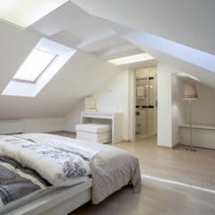 A modern attic bedroom with sloped ceilings and a skylight. The room features a bed with patterned bedding, a small vanity and stool, and an open doorway leading to a bathroom. A floor lamp stands in the corner on light wood flooring.