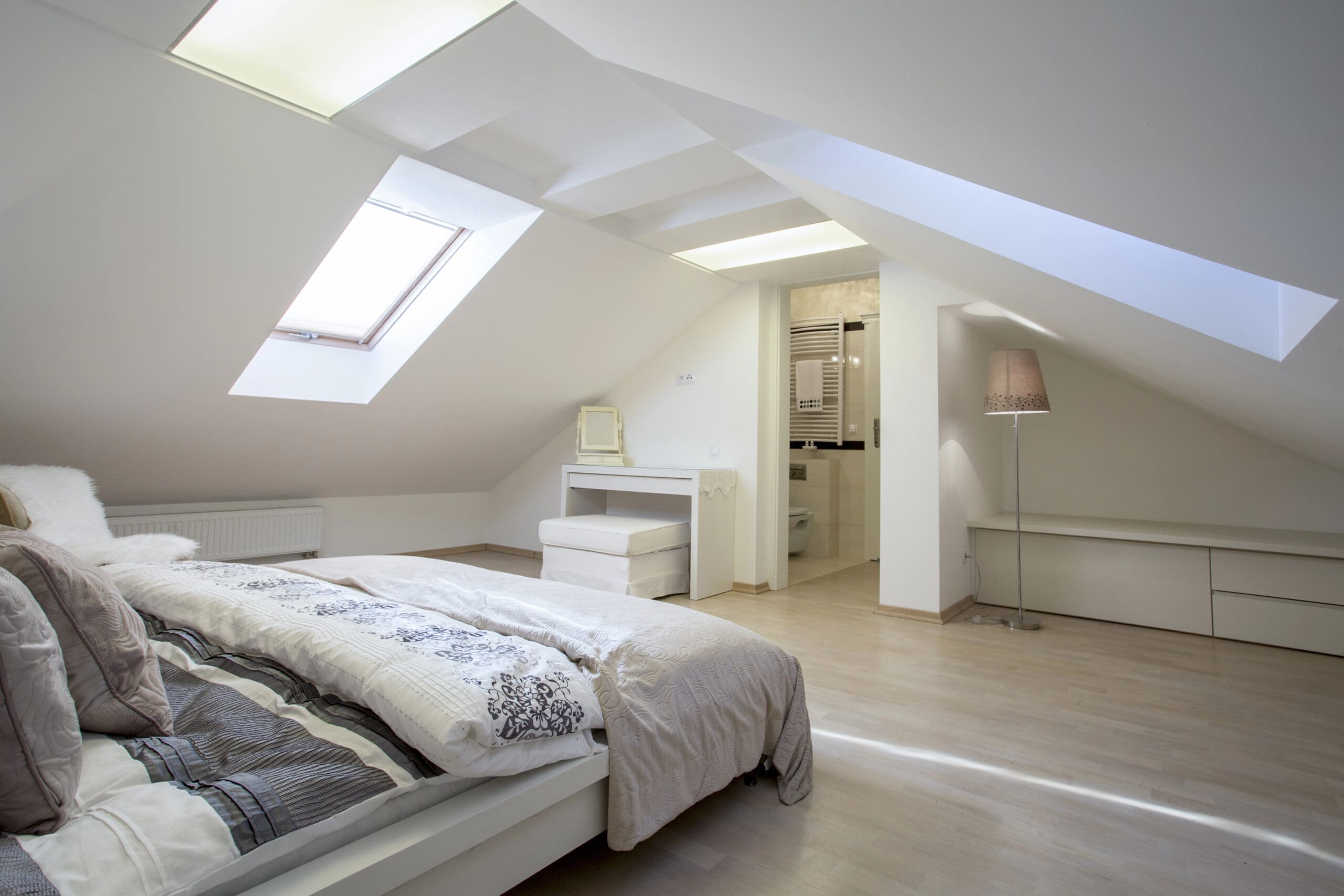 A modern attic bedroom with sloped ceilings and a skylight. The room features a bed with patterned bedding, a small vanity and stool, and an open doorway leading to a bathroom. A floor lamp stands in the corner on light wood flooring.