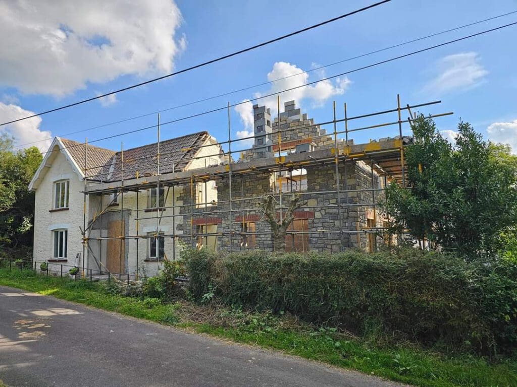 A house under construction with scaffolding on the side. The building has a mix of white and stone walls. It's surrounded by greenery and under a partly cloudy sky. A paved road runs beside the house.
