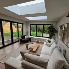 A modern living room with a large skylight and floor-to-ceiling windows, offering a view of lush greenery outside. The room features a light sofa, a wooden coffee table, an armchair, a floor lamp, and decorative wall art.