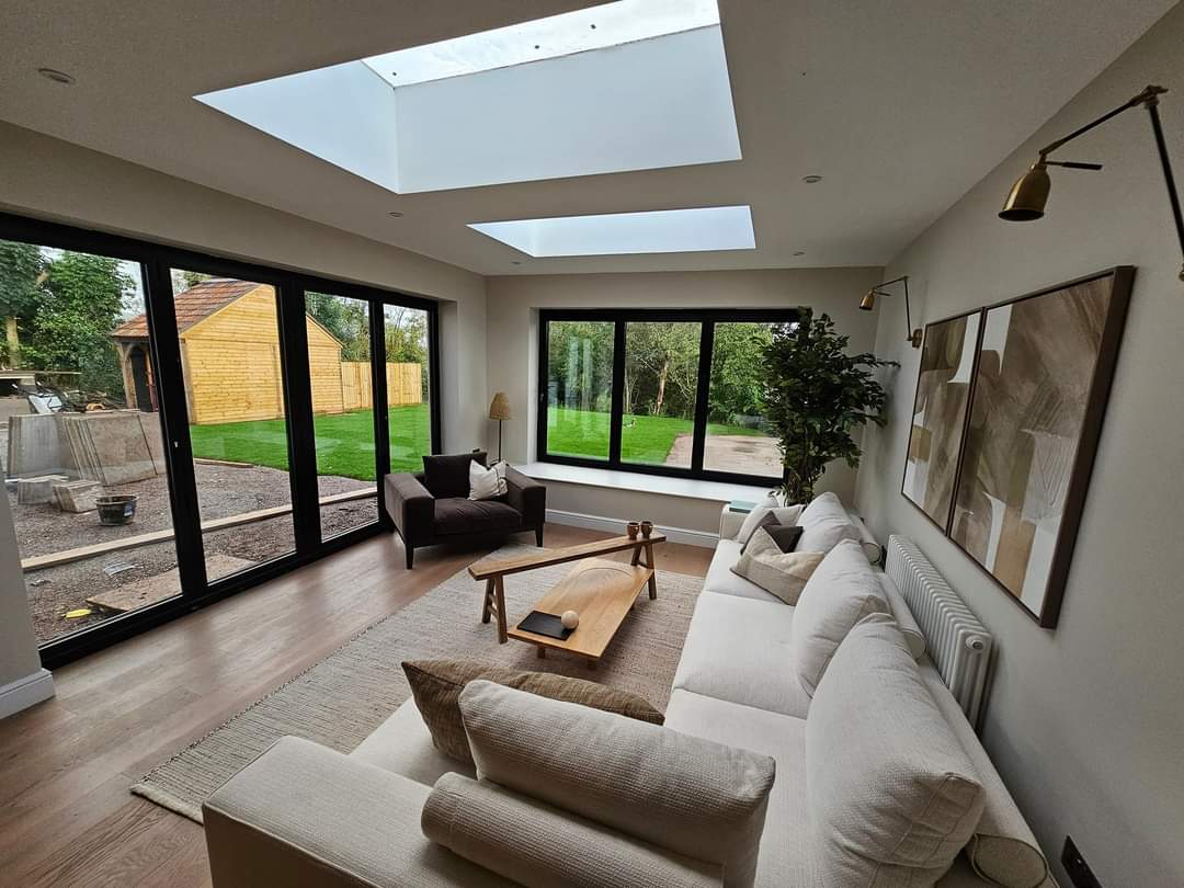 A modern living room with a large skylight and floor-to-ceiling windows, offering a view of lush greenery outside. The room features a light sofa, a wooden coffee table, an armchair, a floor lamp, and decorative wall art.