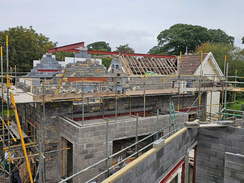 Partially constructed building with stone and concrete blocks. Scaffolding surrounds the site, and a wooden roof frame is visible. Trees and a cloudy sky are in the background.