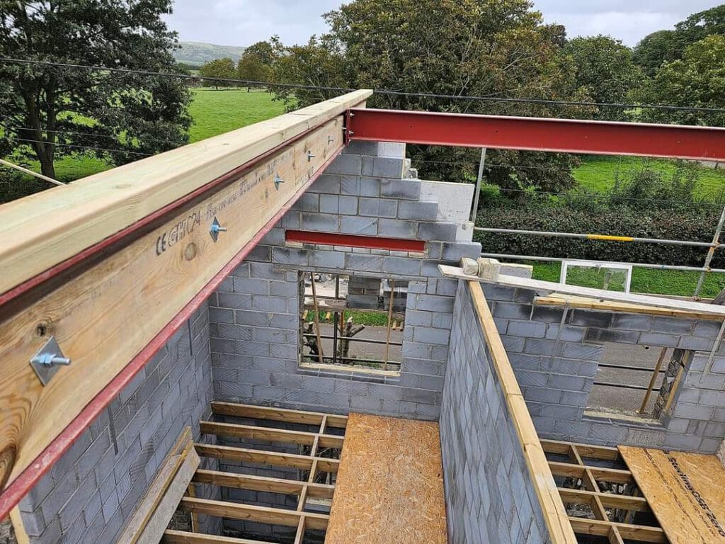 View of a partial building under construction with exposed gray concrete blocks and wooden beams. A countryside landscape featuring trees and green fields is visible in the background.