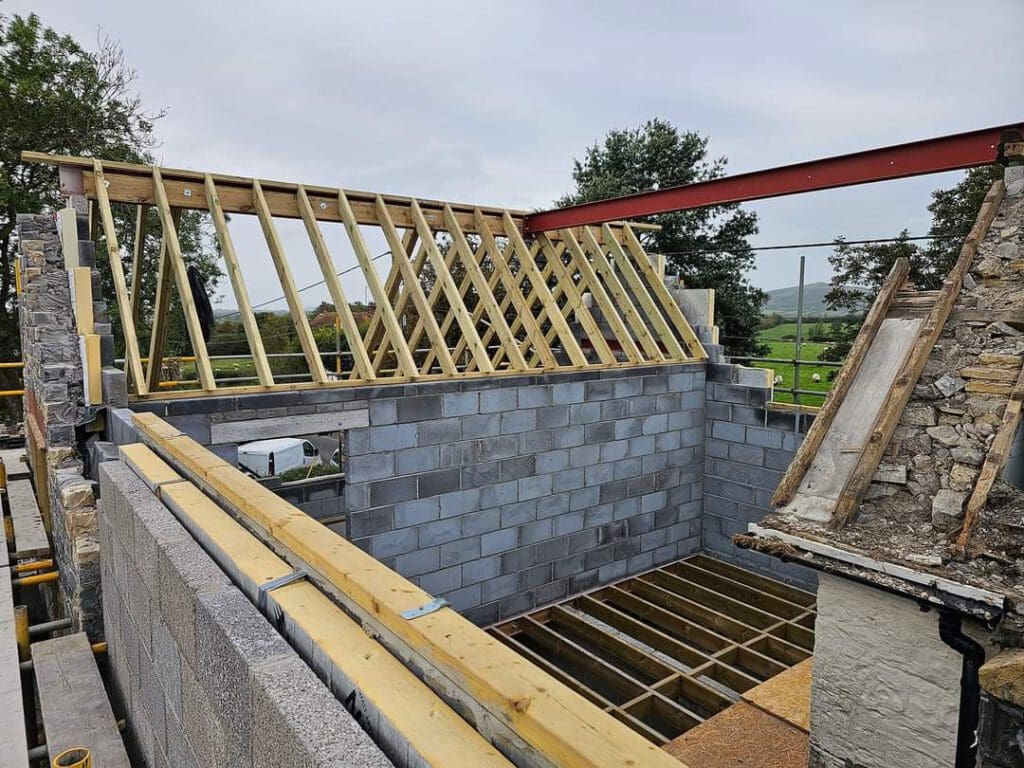 A partially constructed building with gray cinder block walls and wooden roof trusses. The roof is incomplete. Trees and a grassy landscape are visible in the background. Scaffolding surrounds the structure.