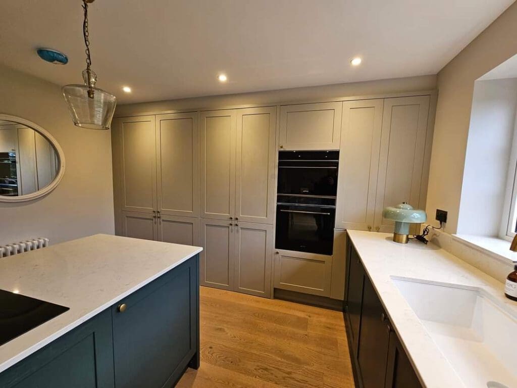Modern kitchen with light gray cabinets, a built-in oven, and a white marble countertop. A blue island with an induction cooktop is centered. A round mirror and hanging light fixture add elegance. Wooden flooring and a window provide a warm atmosphere.