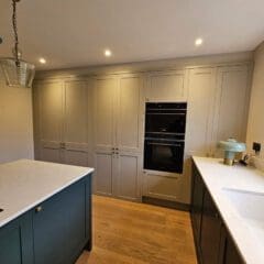 Modern kitchen with light gray cabinets, a built-in oven, and a white marble countertop. A blue island with an induction cooktop is centered. A round mirror and hanging light fixture add elegance. Wooden flooring and a window provide a warm atmosphere.