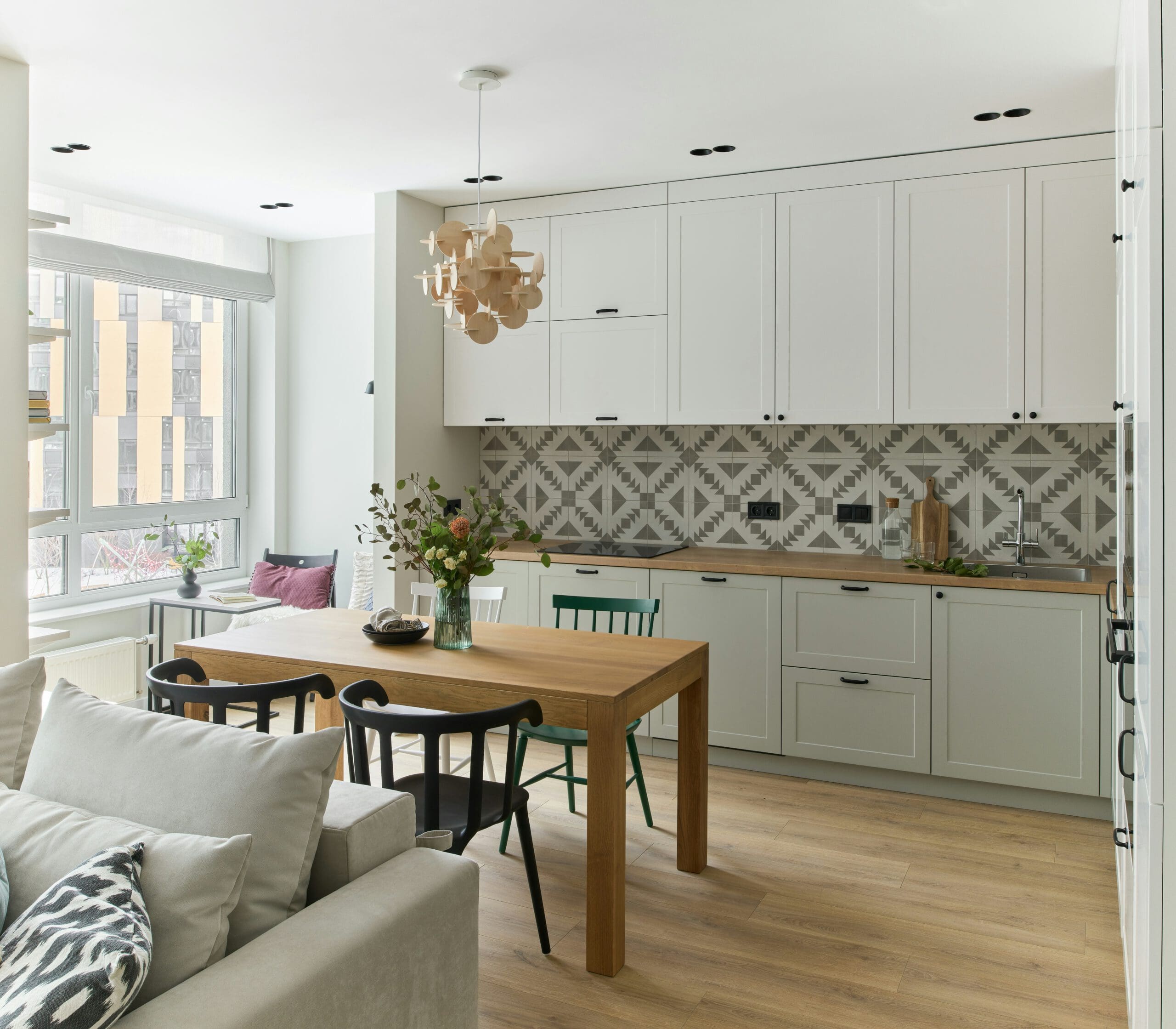 Modern kitchen and dining area featuring white cabinets, geometric backsplash, and a wooden dining table with mixed chairs. A cozy gray sofa and a unique wooden pendant light add warmth. Large windows provide natural light.