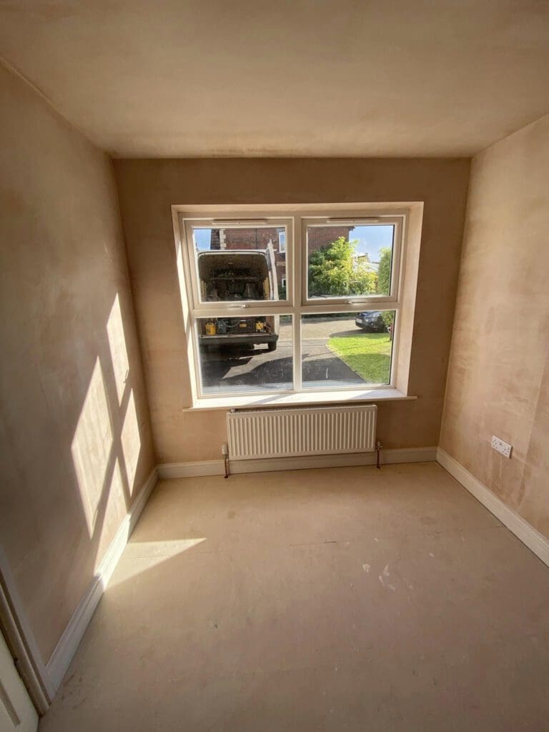 A small, freshly plastered room with sunlight streaming through a double window, casting shadows on the walls. A radiator is positioned under the window, and outside, a truck and a glimpse of a garden are visible.