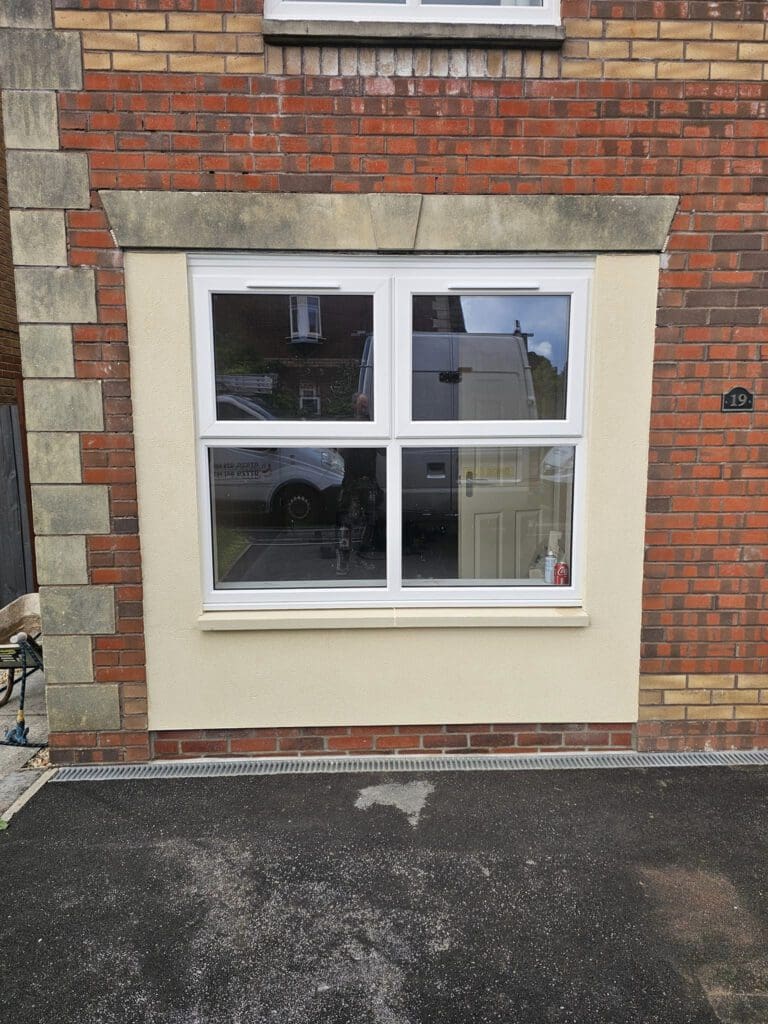 The image shows a rectangular window with four panes set in a brick and plaster wall. The plaster surrounding the window is a light beige color, contrasting with the red brick. A reflection of a van and person is visible in the window.