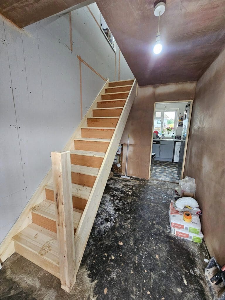 A partially renovated room with bare plastered walls and a concrete floor. An unfinished wooden staircase leads to the upper floor. A lit bulb hangs from the ceiling. Supplies and tools are scattered on the floor, and a doorway opens into a kitchen area.