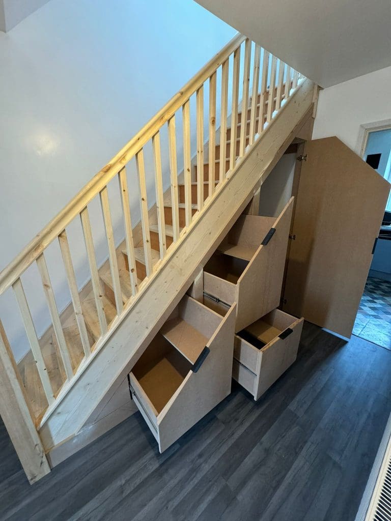 Wooden staircase with a built-in storage unit underneath featuring five pull-out drawers. The space-saving design uses natural wood, and the drawers are open, revealing storage compartments. The hardwood floor complements the light-colored walls.