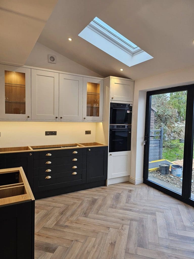 Modern kitchen with white upper cabinets and black lower cabinets. Built-in double oven, wood countertops, and glass display sections. Skylight above and large glass doors leading to a garden. Herringbone-patterned wooden floor.