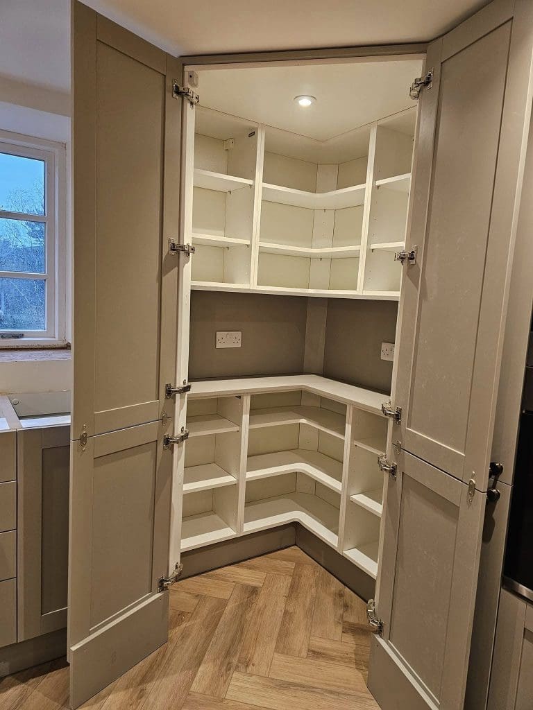 Open corner kitchen pantry cabinet with grey doors, revealing multiple white shelves inside. The shelves form a U-shape to fit the corner space. The herringbone-patterned wooden floor adds a warm touch beneath the cabinet.