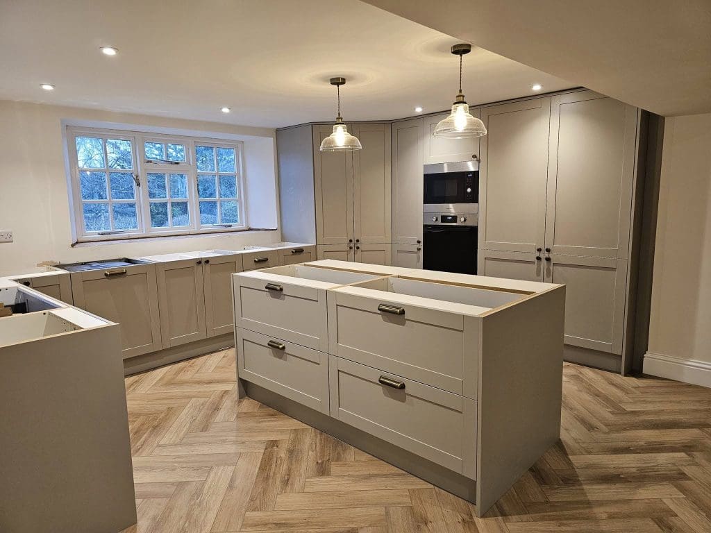 A modern kitchen with light gray cabinets and a large central island with drawers. Two pendant lights hang above the island. The room features herringbone wood flooring and a window showing trees outside. Built-in oven and microwave are visible.