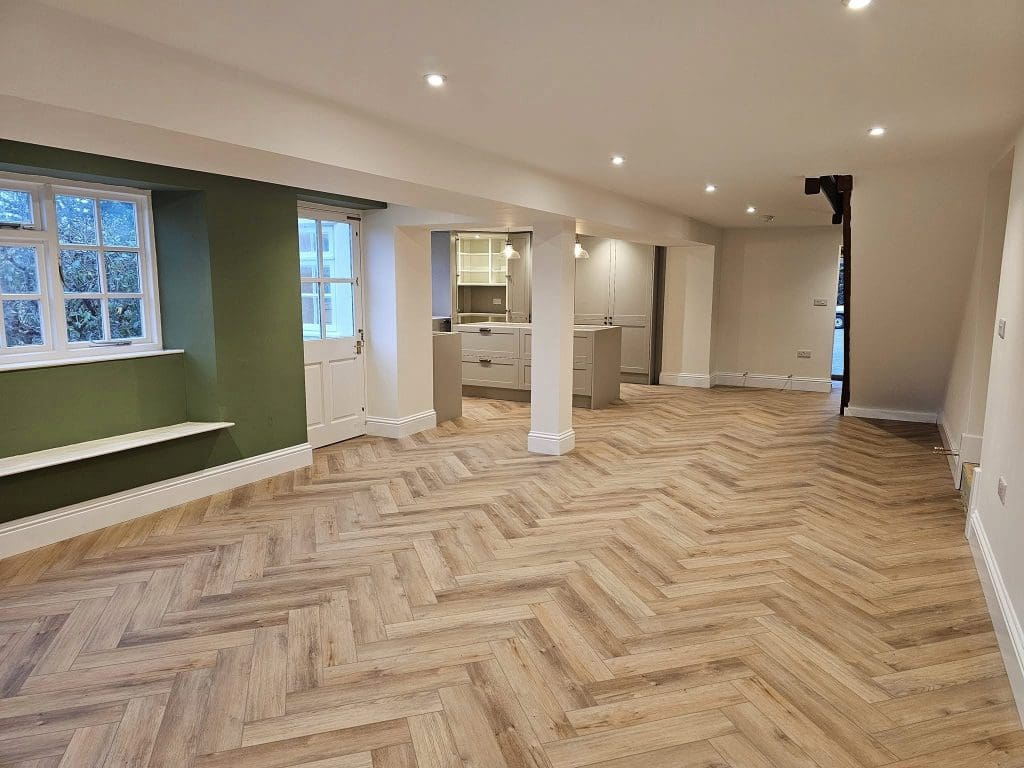 A spacious room with herringbone-patterned wooden flooring and white walls. There are large windows and a door on the left, a green accent wall with a bench, and an open kitchen with white cabinets in the background. Ceiling lights are on.