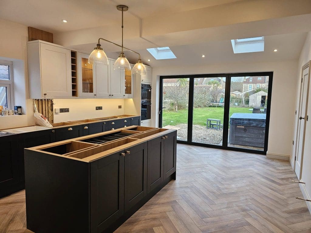 Modern kitchen with black and white cabinetry, pendant lights, and herringbone-patterned wooden floor. Large windows offer a view of a spacious garden. An island unit is centered without a countertop installed.