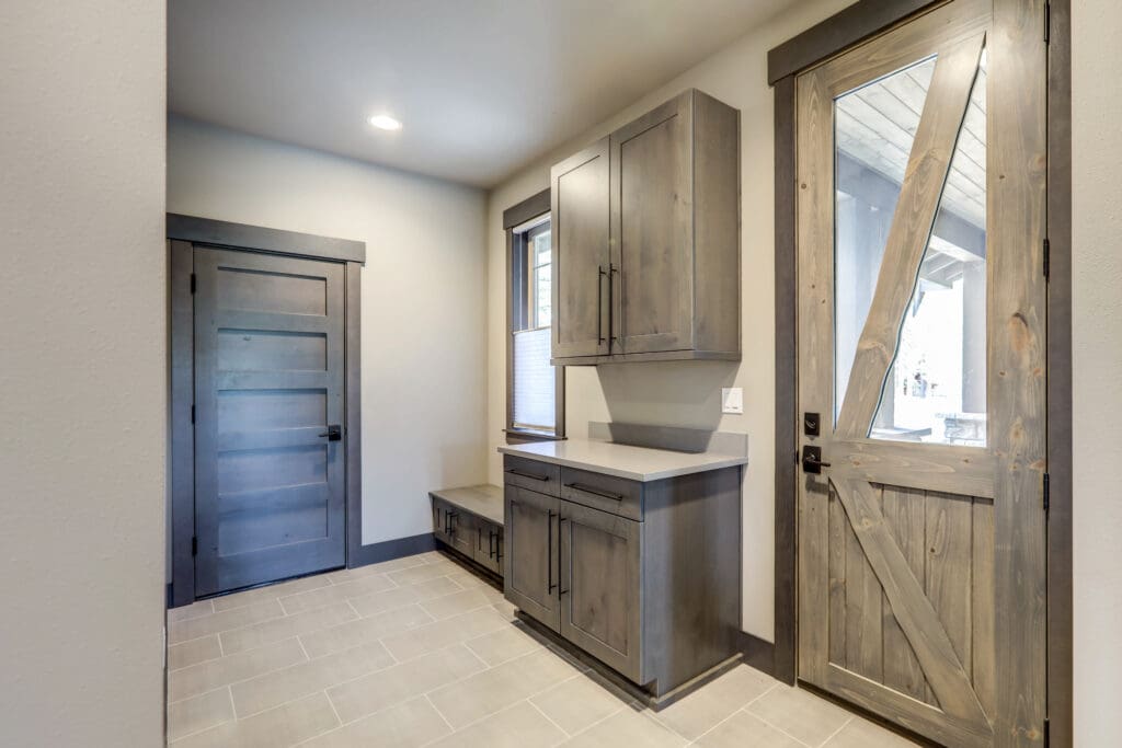 A modern mudroom featuring light tiled flooring and wooden cabinetry with a built-in bench. There are two rustic wooden doors and a window, creating a cozy and practical entrance area.
