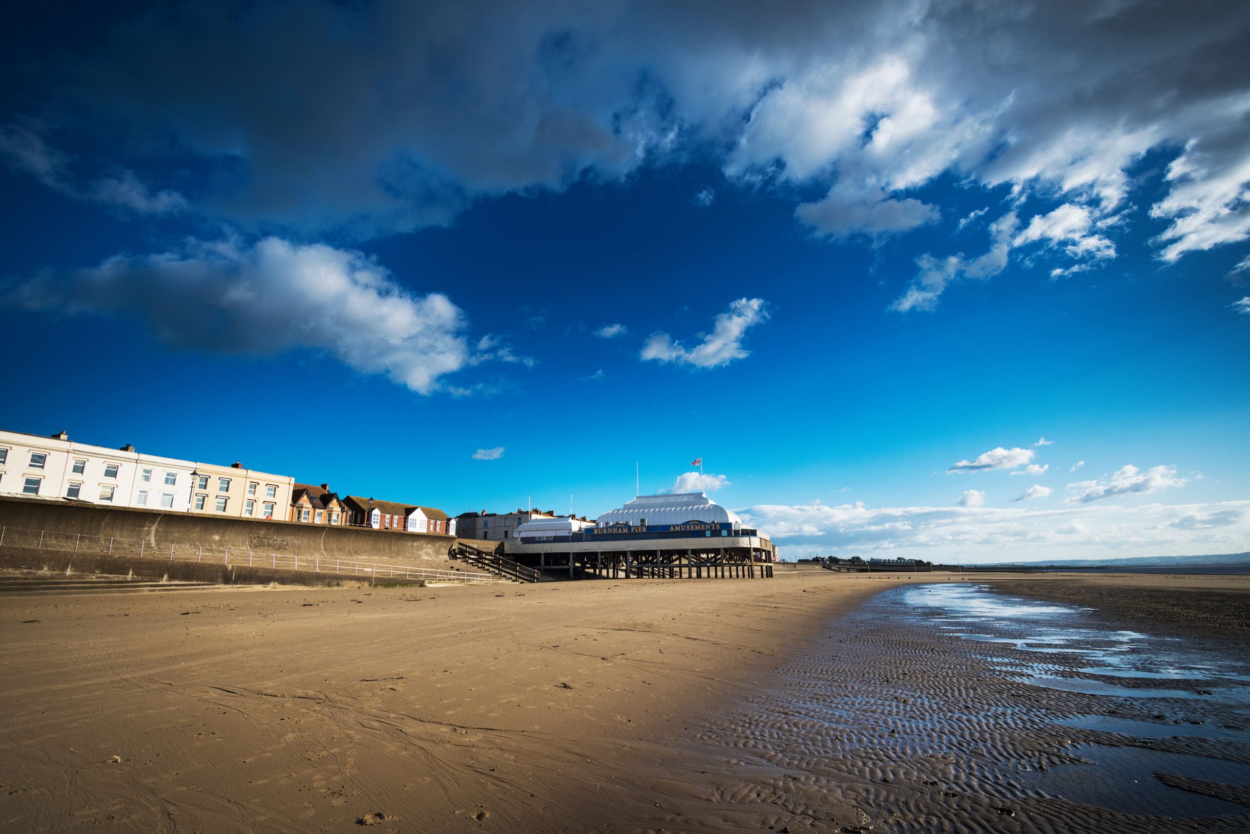 A wide sandy beach under a bright blue sky with some clouds. On the left, a row of colorful buildings lines the shore. In the center, a white pier extends over the sand. The tide is low, leaving wet ripples on the sand.