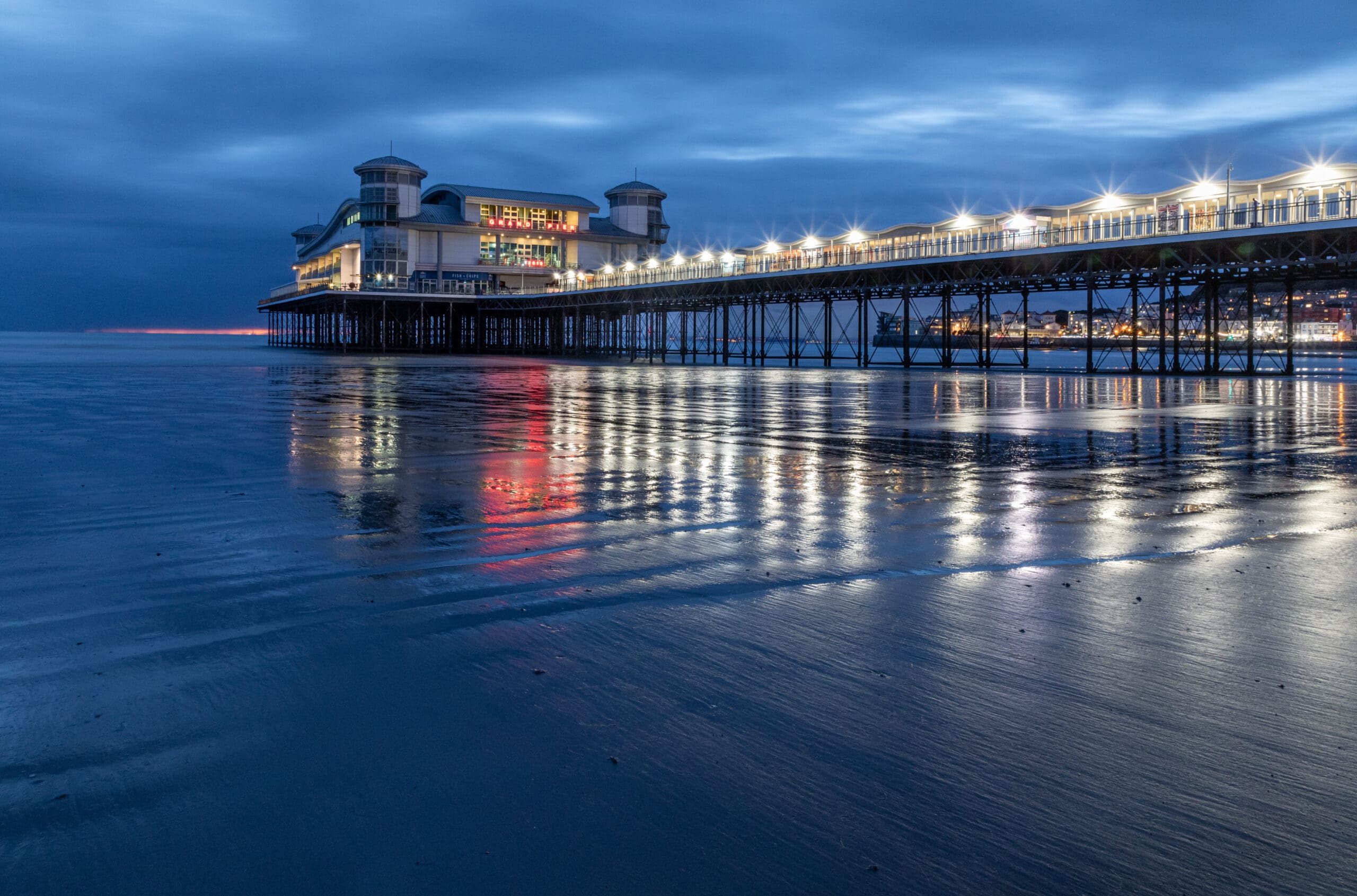 A pier lit with bright lights stretches into the evening sky over calm waters. Reflections ripple on the wet sand and ocean below. The background features a large building at the pier's end, with a vibrant, illuminated facade.