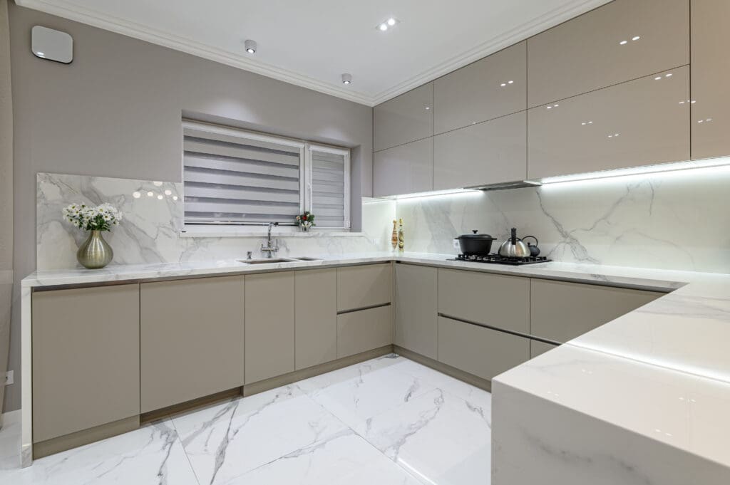 Modern kitchen with beige cabinets, marble countertops and backsplash, and polished white marble floor. A stainless steel sink is by the window with striped blinds. A vase with flowers is on the counter. Built-in lights illuminate the space.