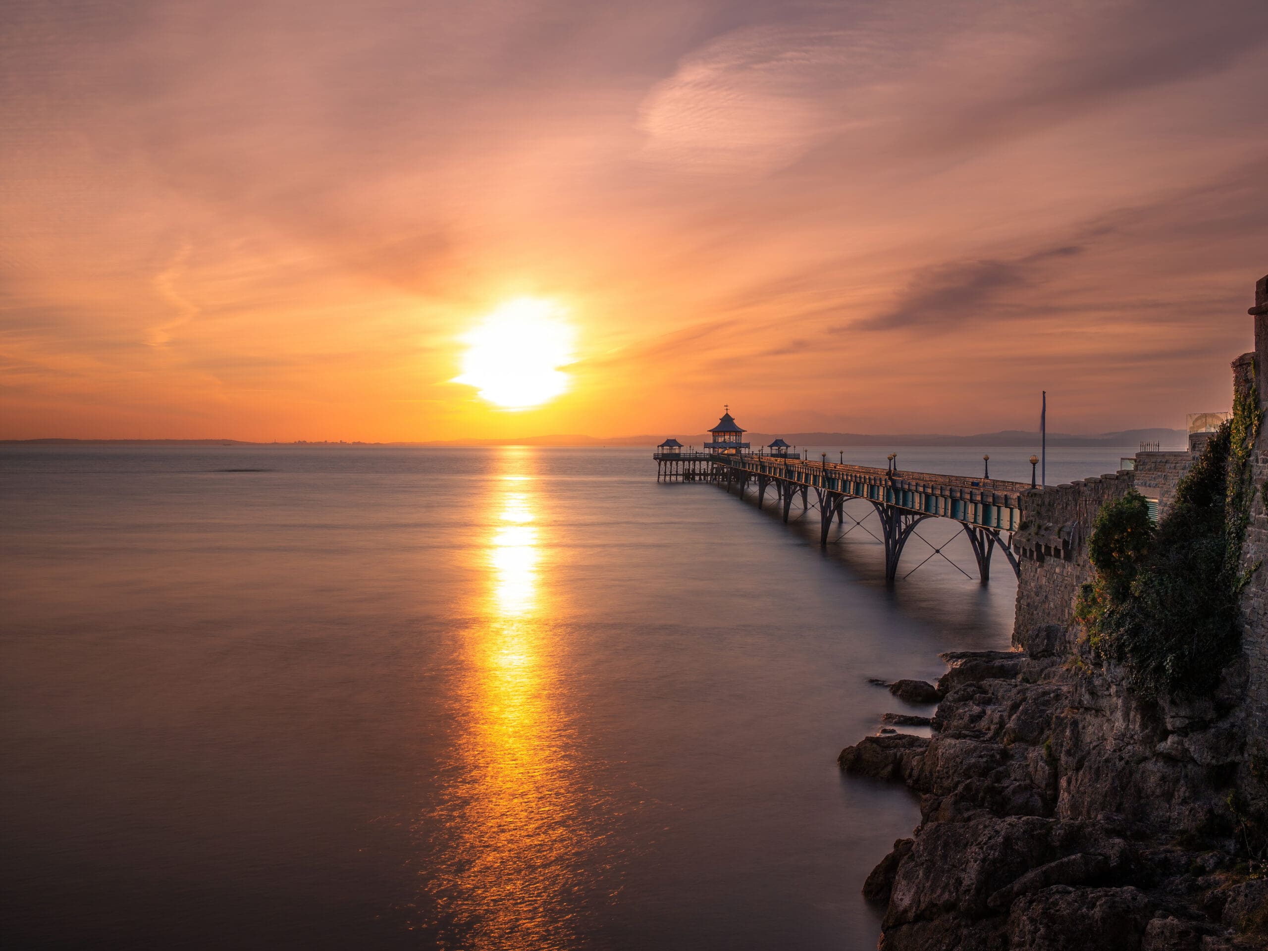 A scenic view of a long pier extending into the sea at sunset, casting a shimmering reflection on the calm water. The sky is painted with warm orange and pink hues, and rocky cliffs are visible in the foreground.