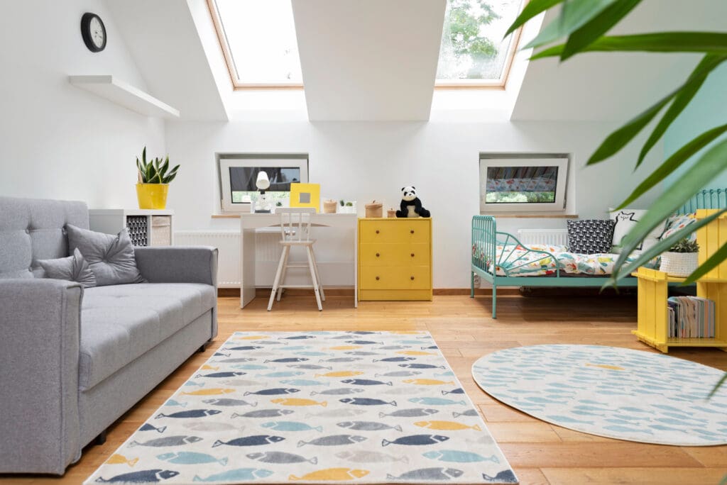 A cozy bedroom with a gray sofa, a colorful rug with fish patterns, a small bed with a blue frame, and a study area equipped with a desk and chair. Skylights and small windows allow natural light, and plants add a touch of greenery.
