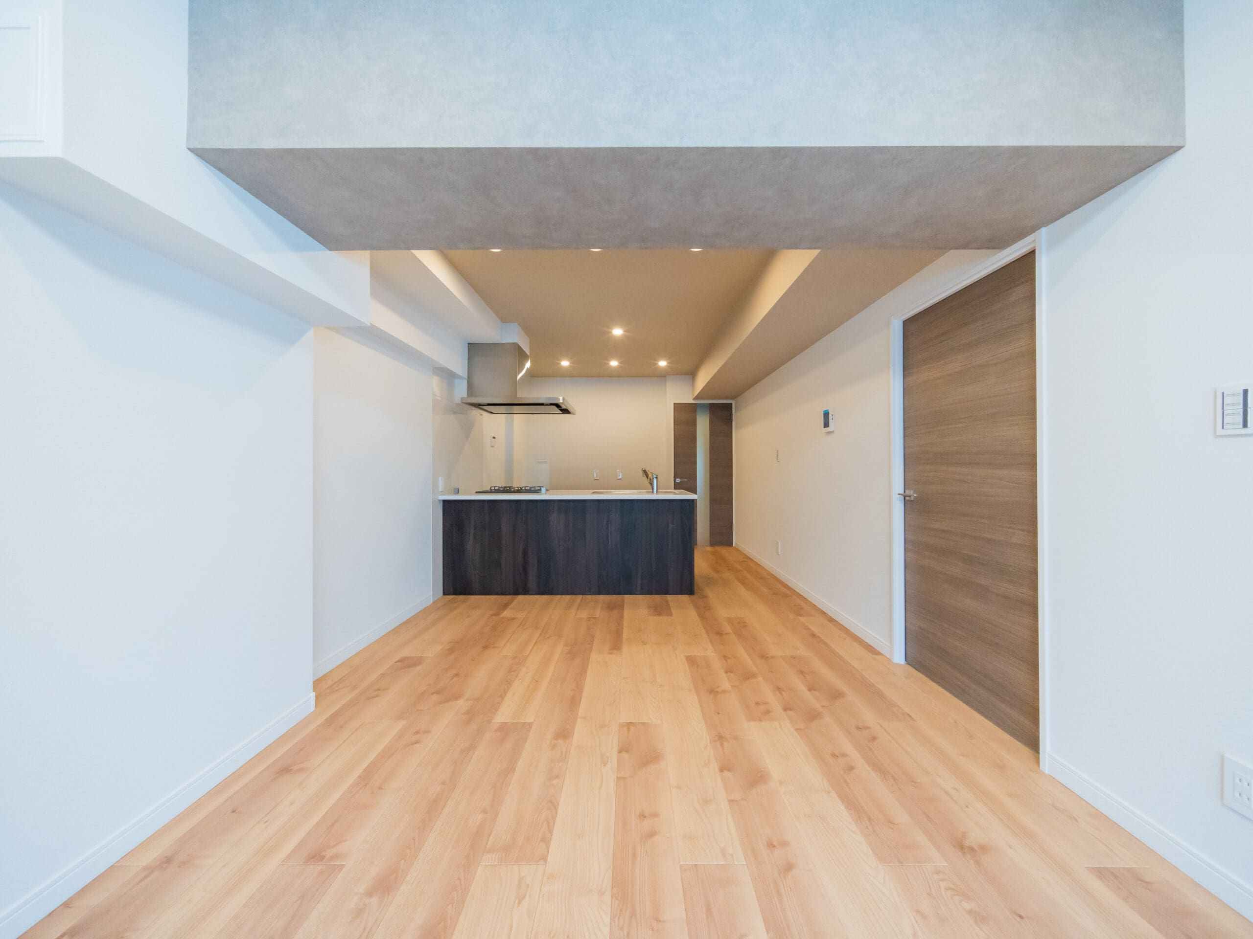 Modern apartment interior with wooden flooring, light-colored walls, and a recessed ceiling. The space features a small kitchen area with dark cabinetry and built-in lighting. Two wooden doors are visible on the right side.