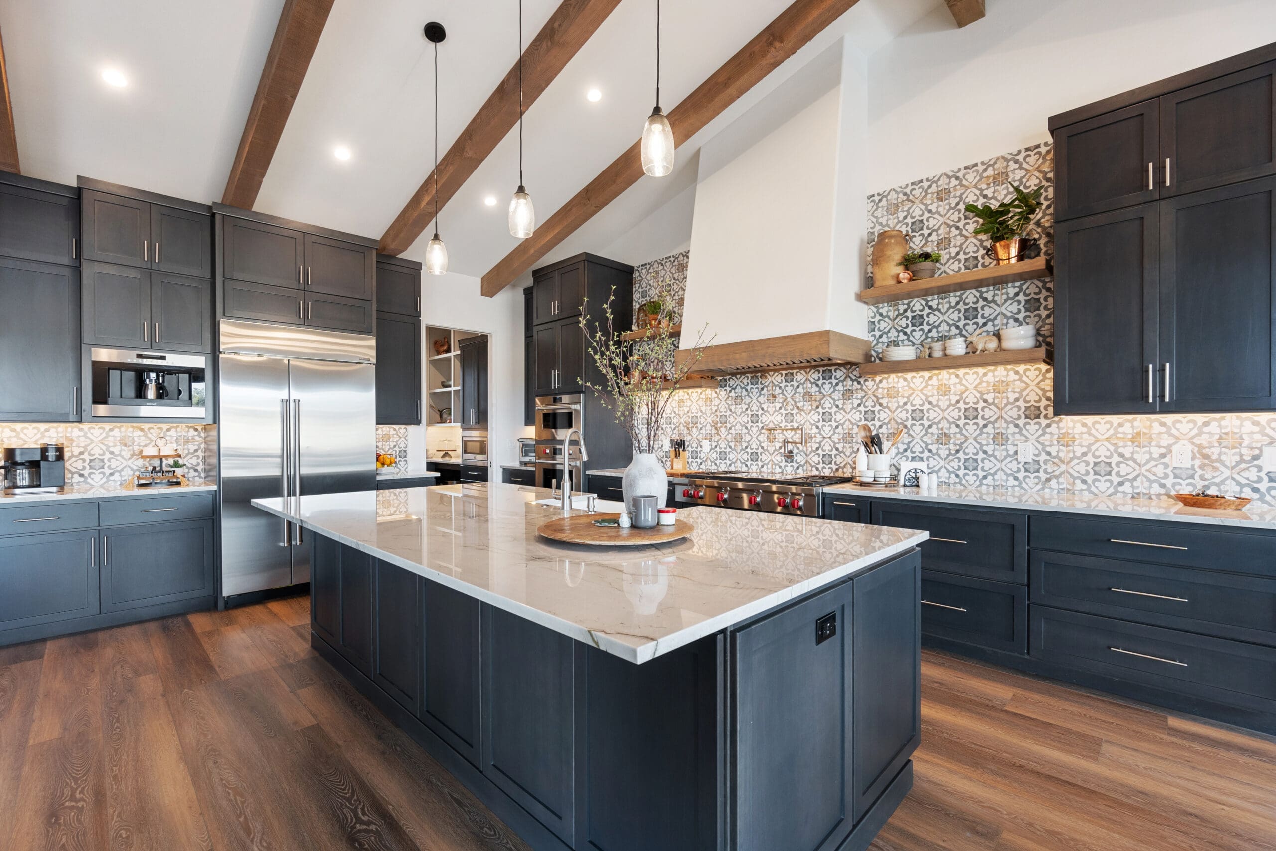 A modern kitchen with dark cabinets, a large island with a white countertop, and wooden ceiling beams. The backsplash features decorative tiles, and pendant lights hang above the island. Stainless steel appliances are visible throughout.