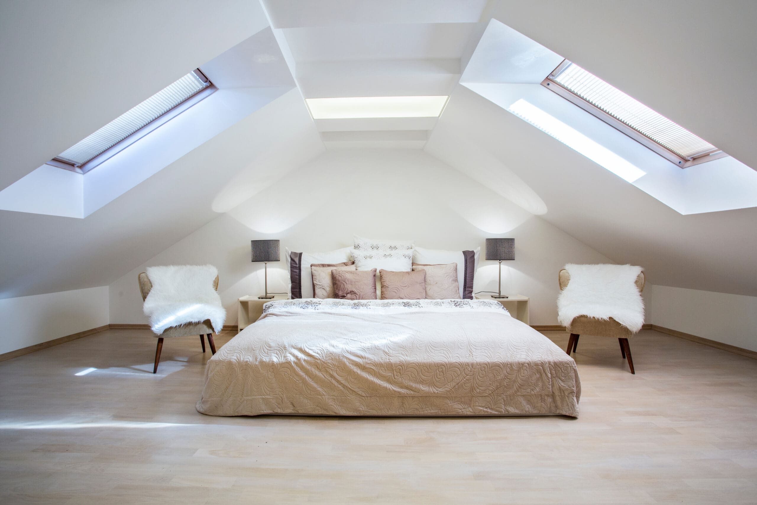 A cozy attic bedroom with slanted ceilings features a large bed with beige bedding, flanked by two nightstands with lamps. Two chairs with white faux fur throws stand on either side. Natural light comes in through skylights overhead.