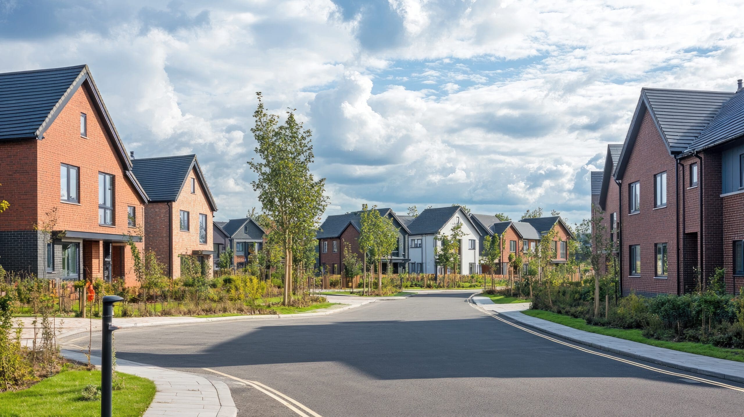 A suburban street with neatly lined modern brick houses, small trees, and landscaped lawns under a partly cloudy sky. The road curves gently, and the scene is calm and orderly.