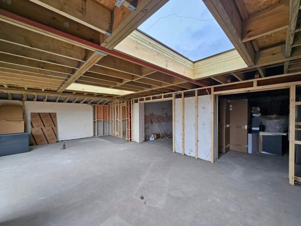 An unfinished room with exposed wooden ceiling beams and a large square skylight. Cardboard boxes and building materials are stored along one wall. The floor is concrete, and some walls are partially constructed.