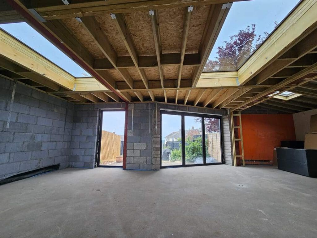 Interior view of a building under construction with exposed wooden beams and unfinished walls. Large sliding glass doors open to a garden with trees. Sunlight enters through a skylight, illuminating the concrete floor.