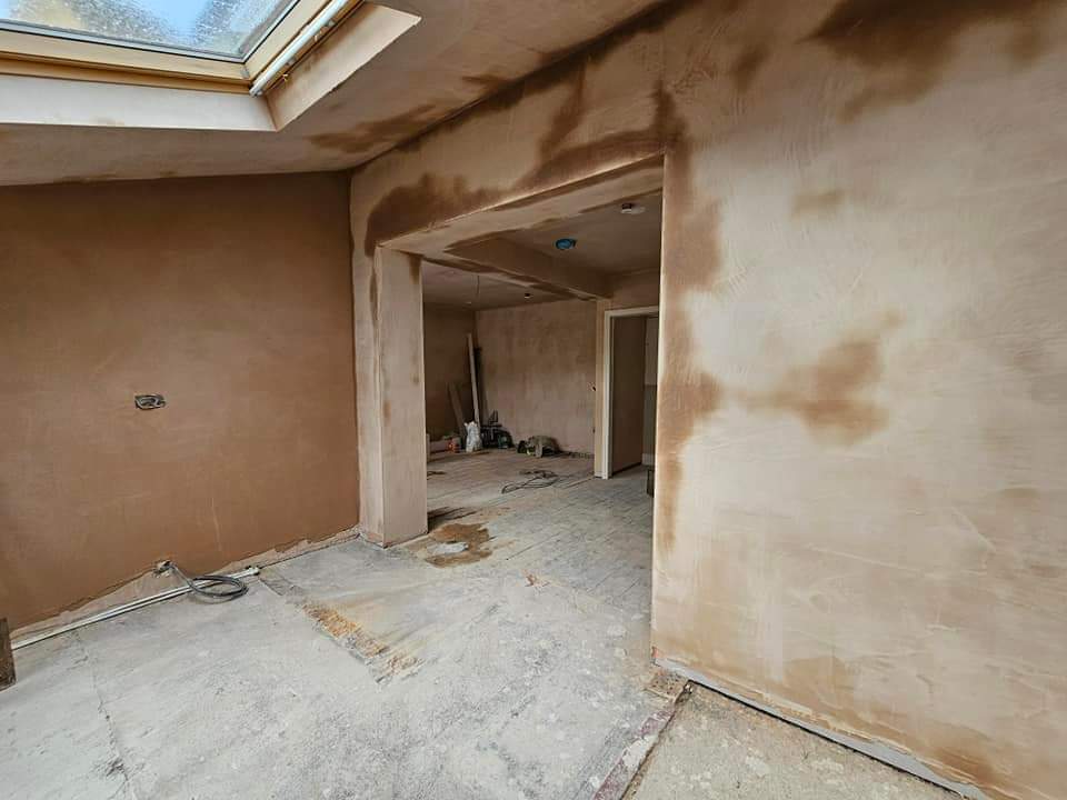 A room under construction with freshly plastered walls. There is a skylight above, and tools are scattered on the unfinished floor. An open doorway leads to another area.