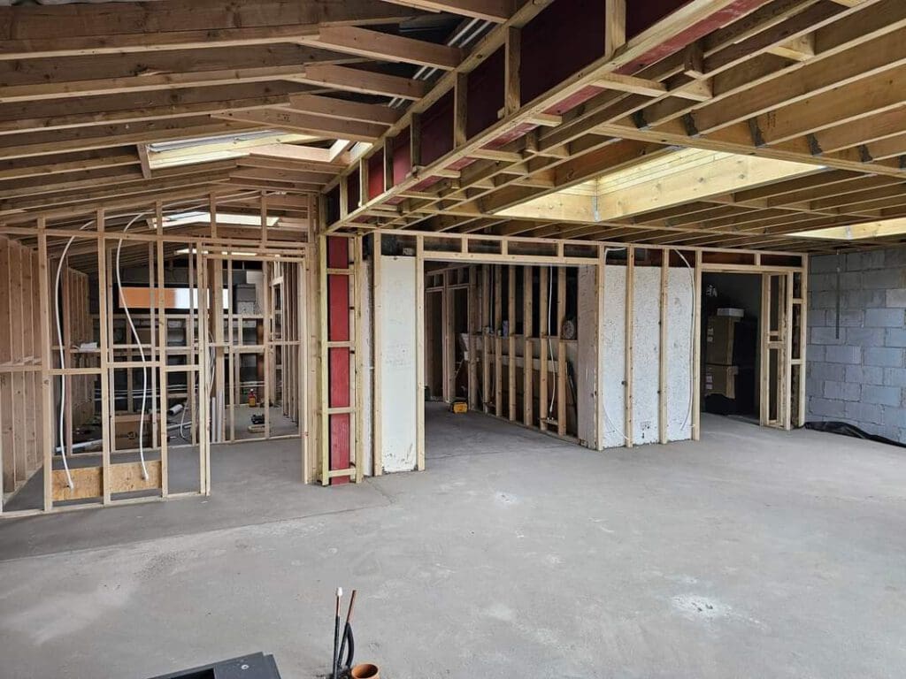 Interior of a partially constructed building with exposed wooden framing and beams. The floor is bare concrete, and there are openings for windows and doors. The ceiling shows unfinished wood and insulation.