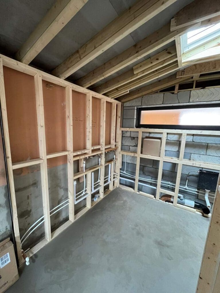 A room under construction with exposed wooden framing and pipes on the walls. There is a window with frosted glass on the right wall and a skylight on the slanted ceiling. The floor is unfinished concrete.
