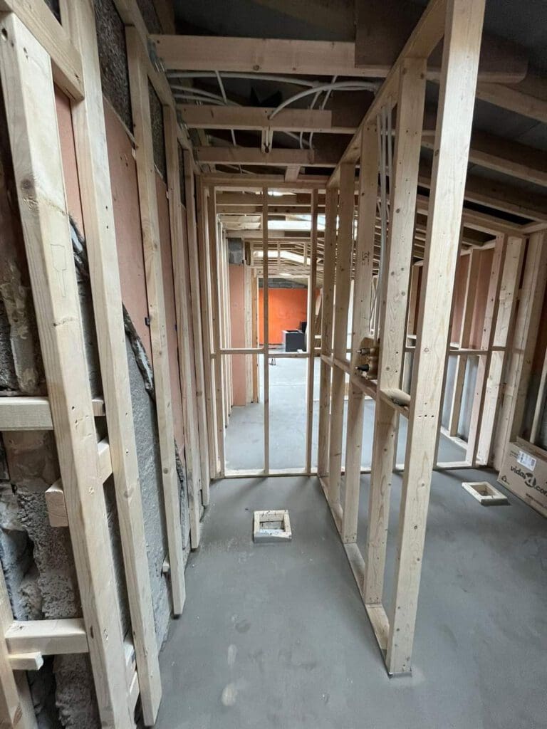 View of a building under construction, featuring wooden framing and exposed beams. The structure reveals interior walls and pathways in progress, with an unfinished floor and ceiling in the background. Natural light enters from an open space ahead.
