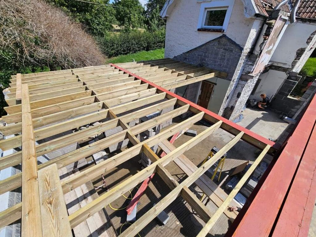 An overhead view of a house extension under construction. Wooden beams form a new roof structure beside the existing building. A person is sitting in the open area under the extension's framework. Nearby trees and bushes are visible.