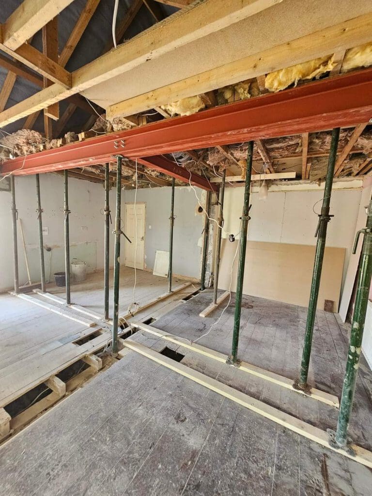 Construction site inside a building with an exposed ceiling and walls. There are metal support beams and temporary scaffolding. Some insulation is visible in the ceiling. The floor is partially covered with wooden planks.