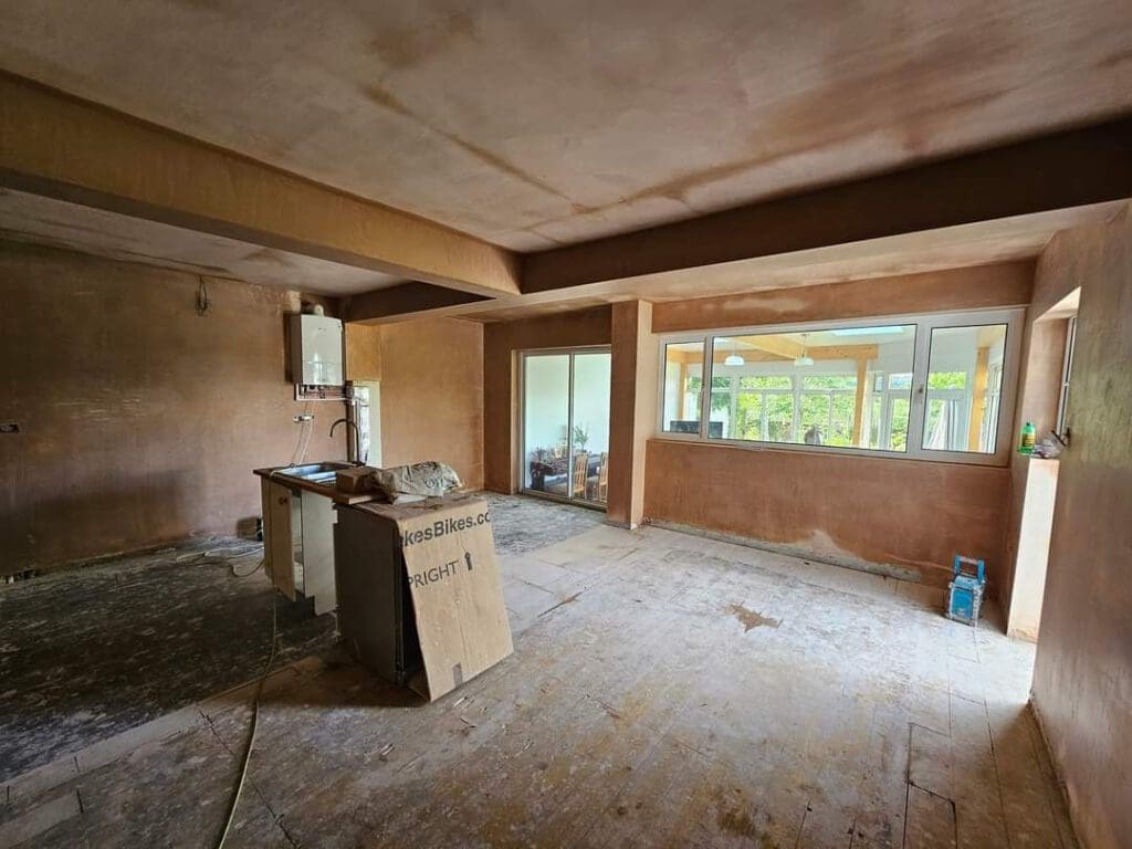 A spacious, unfurnished room with bare plastered walls and a wooden floor. Large windows and sliding doors reveal a view of greenery outside. A makeshift table holds a cardboard box and some items are scattered on the floor.