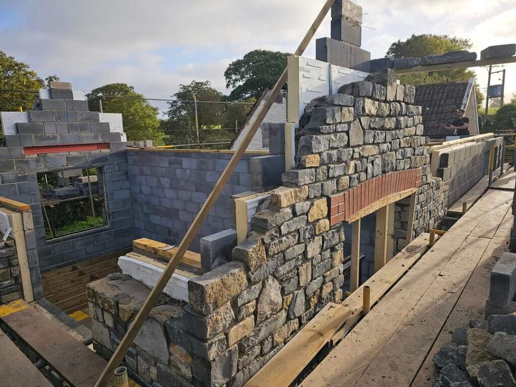 A partially constructed building with stone and brick walls is surrounded by scaffolding. The sky is partly cloudy, and trees are visible in the background. A person is standing on the scaffolding to the right.
