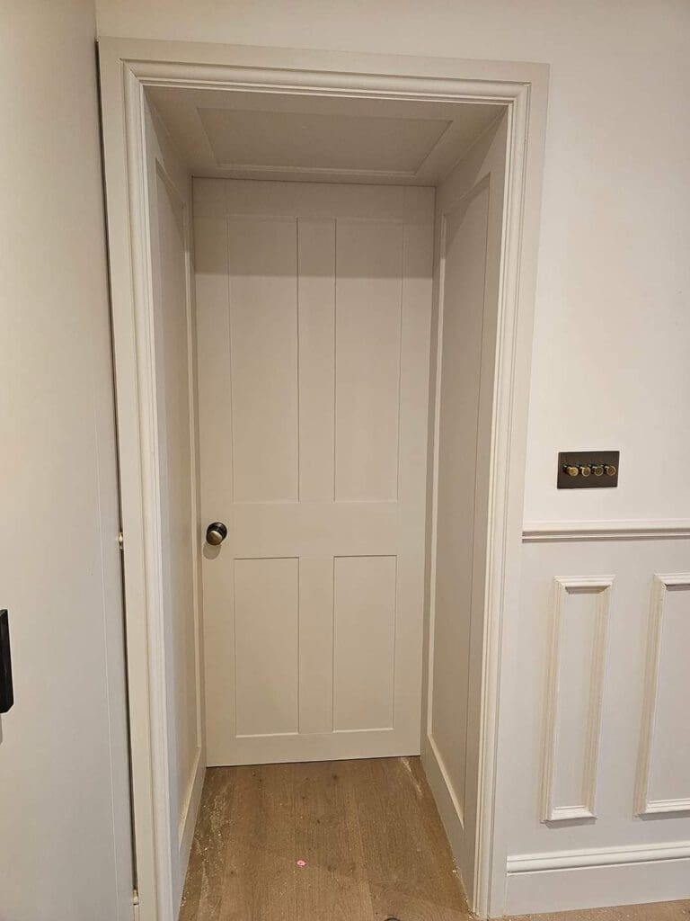 A closed white door with a simple panel design, set in a white wall. There is a dark metal doorknob on the left, and a black light switch with multiple buttons on the right. The wooden floor is light-colored.