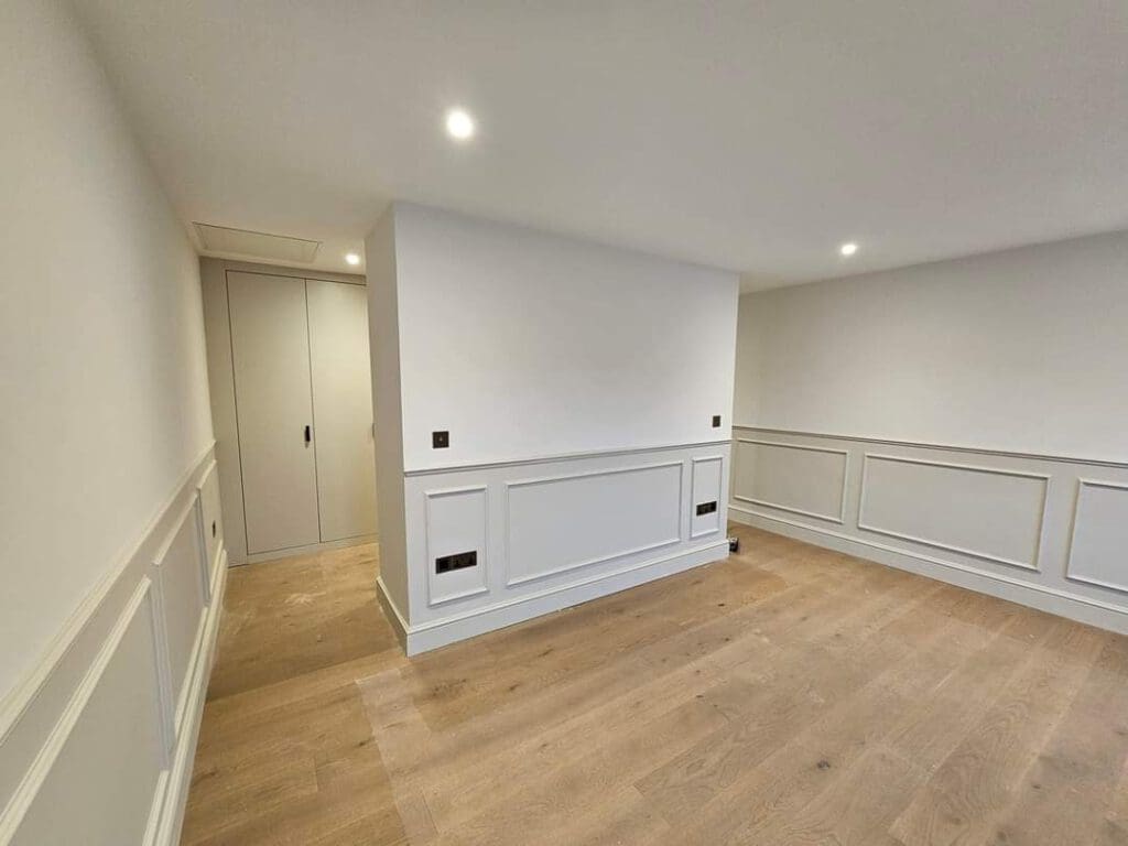 An empty room with light wood flooring, white walls, and recessed lighting. It features wainscoting panels and a small hallway leading to a closed door. The ceiling is white, enhancing the room's brightness.
