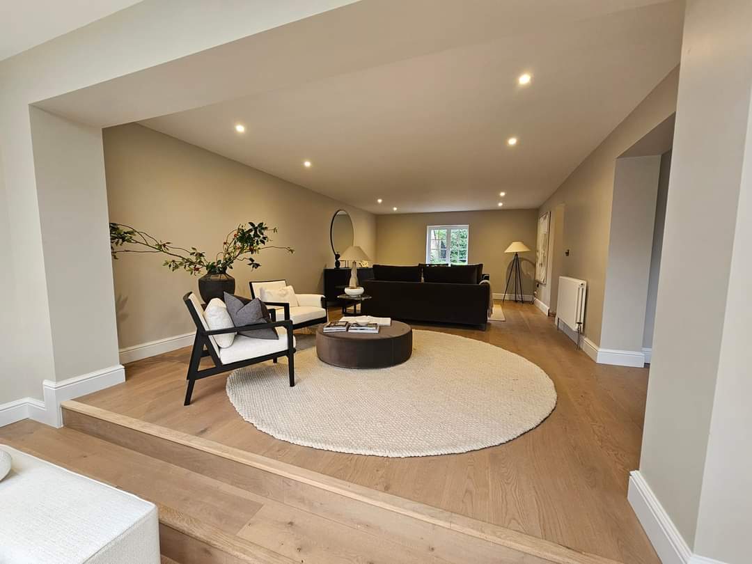 A modern living room with beige walls and wooden flooring. It features a black sofa, a round beige rug, black and white chairs, a coffee table, a tall lamp, and a plant in a vase. Recessed lighting brightens the space.