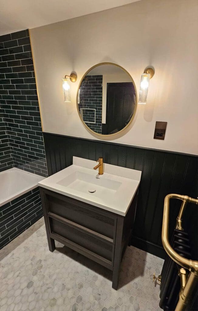 A modern bathroom features a circular mirror with two vertical lights on either side, above a dark vanity with a white countertop and gold faucet. The walls have dark green tiles and wainscoting, and the floor is covered with hexagonal tiles.