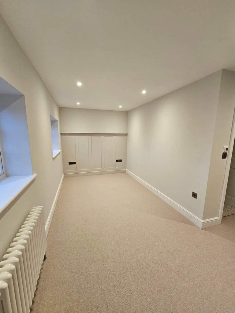 A minimalistic room with beige carpet, light gray walls, and recessed ceiling lights. There's a radiator under a window on the left and built-in white cabinets at the end of the room. A doorway is visible on the right.