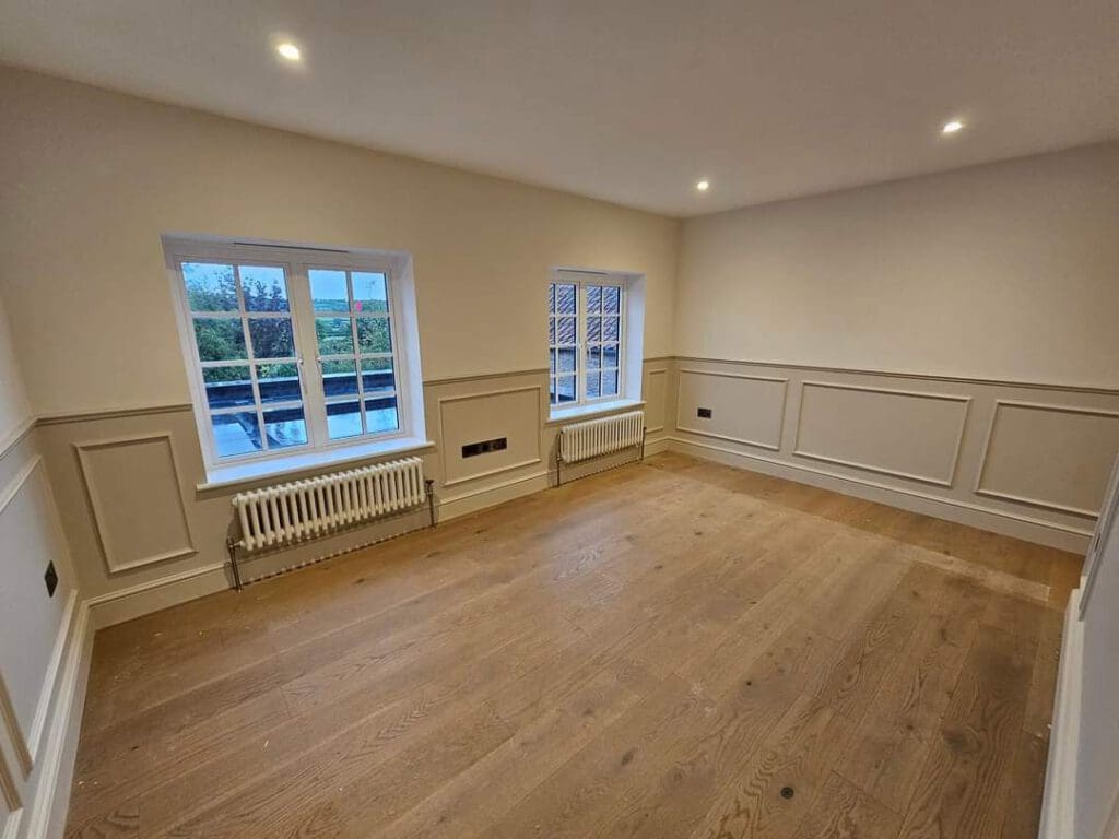 A spacious, unfurnished room with light-colored walls and wooden flooring. Two windows with grid patterns let in natural light. The walls feature decorative paneling, and radiators are located beneath the windows. Recessed lighting is installed in the ceiling.