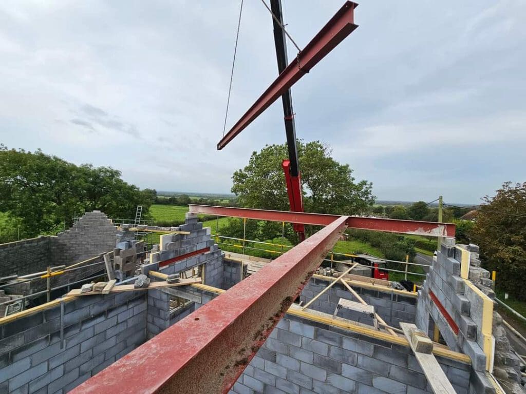 Construction site view from above, showing a building in progress with grey cinder block walls. A large red steel beam is being installed with a crane. Green trees and open fields are visible in the background under a cloudy sky.