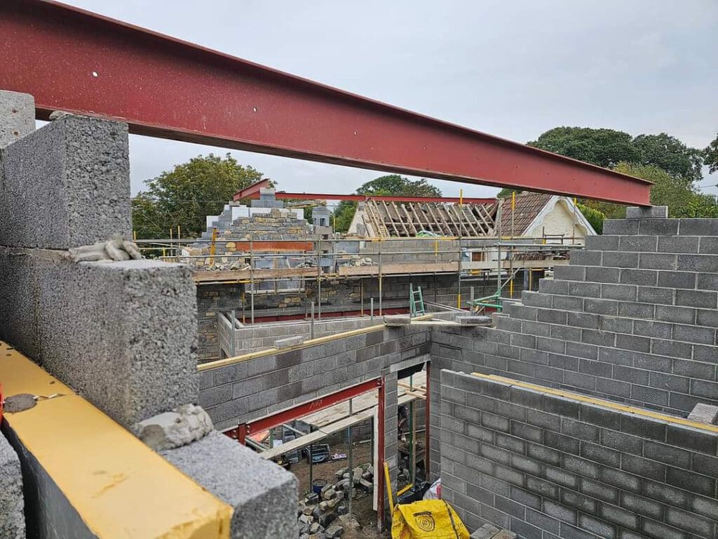 Construction site with partially built brick walls and exposed steel beams. Scaffolding surrounds the structure, and building materials like cement blocks are visible. Trees and parts of nearby houses can be seen in the background.